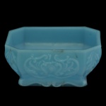 Tigela em vidro opalinado de cor azul. Apresenta um motivo floral em relevo e sustentada por uma base retangular lisa. 6 x 9 x 12 cm