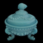 Bomboniere circular funda em vidro opalinado na cor azul moldada e ornada por frisos estilizados e perolados. Recipiente ladeado por largas pernas recurvas com pés revirados. Tampa abobadada, seguindo a decoração e encimada por pega em botão floral. Sob a base pequeno adorno no formato de rosácea.18 x 14 cm.24 x 14 cm.