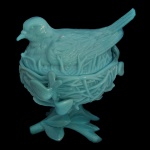Manteigueira em vidro opalinado de cor azul. Apresentando um pássaro sentado sobre um ninho detalhado conhecido como "Bird on Nest" (Pássaro no Ninho). O design em duas partes inclui o ninho como base e o pássaro como tampa, com detalhes minuciosos nas penas e nos ramos do ninho. 16 x 15 x 13 cm.