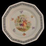 SCHUMANN, Bavaria - Prato em porcelana alemã.  Apresenta uma cena galante romântica de época no centro e borda rendada fenestrada. Marca da manufatura na base. 19 cm.