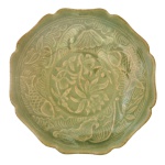 DINASTIA MING (1368-1644). SÉC. XVII - Delicado bowl raso em porcelana chinesa, de formato lobulado, com borda ondulada e oitavada. Apresenta coberta celadon do tipo Yaozhou ou celadon do Norte, levemente craquelada, e ornamentada por adornos incisos. Margem decorada por carpas e lotus, centralizando medalhão floral. Base baixa e retraída.