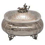 Bomboniere de formato bojudo, oval e achatada, em prata brasileira, repuxada e cinzelada. Apresenta decoração com reservas emolduradas por folhagens e volutas, alternadas por faixas florais. Tampa levemente abobadada, acompanhando o estilo, encimada por pega no feitio de anjinho. Pés vazados e revirados. Contrastes do teor de 833 mls e marca do fabricante Pratalei. 16 x 20 x 17 cm. Peso 823 gramas.