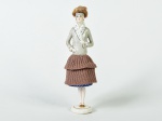 Boneca em porcelana esmaltada e vitrificada, saia em tecido na cor marrom. Altura: 19,5cm.
