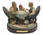 ATHENA COLLECTION - Grupo escultórico com a representação de um casal de idosos degustando chá à mesa; em resina multicolorida sobre base oval de madeira. Medidas: 20 x 15 cm. Base com ínfimos desgastes na pintura.