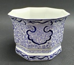 Cachepot octogonal adornado com volutas, elementos geométricos e motivos florais e vegetalistas. Em porcelana decorada à mão na cor azul cobalto. Assinatura da artista na base. Medidas: 17 x 12 cm.
