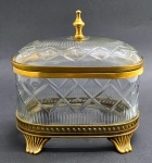 Antigo potiche oval em cristal translúcido, com tampa articulada e estruturado em bronze; cristal lapidado com elementos geométricos e estelares; apoiado sobre quatro pés em forma de volutas. Com inscrição em algarismos indo-arábicos gravada na base. Medidas: 20 x 13 x 19 cm.