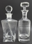 02 licoreiras em cristal translúcido, com diferentes formatos; licoreira com tampa globular com corpo retangular moldado com efeito torcido; licoreira com tampa em forma de disco com corpo cilíndrico lapidado com dois frisos centrais. Medidas: 29 x 13 cm;. 29 x 9 cm. Tampa globular com bicado na base.