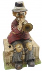 SEIJI NAKANE, MELODY IN MOTION - Grupo escultórico com a representação de um palhaço sentado em caixote de madeira e tocando trompete; caixa de música acoplada. Em porcelana policromada, fabricado no Japão. Medidas: 30 x 17 x 16 cm. Não testado.