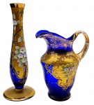 01 solifleur e 01 jarra em vidro azul cobalto, adornado à mão no estilo Rococó com volutas, arabescos e medalhões dourados, e coloridos motivos florais com pétalas em relevo. Solifleur com selo de procedência (Bohemia, antiga Tchecoslováquia). Medidas: 20,5 x 7,5 cm; 13 x 14,5 cm. Jarra com douração desvanecida em alguns locais.