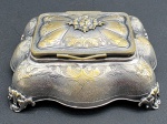 Antiga caixa de jóias em metal espessurado à prata, decorada no estilo clássico com padrões fitomórficos e florais em relevo; tampa articulada; corpo moldado com ondulados e base com quatro pés em forma de volutas. Medidas: 18 x 14 x 7 cm. Com desgastes no banho.