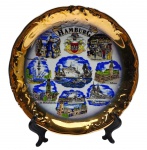 PETER MENK SOUVENIRS - Prato decorativo souvenir da cidade de Hamburgo (Hamburg), Alemanha; estampado com brasão de armas e pontos turísticos da cidade; em porcelana multicolorida, com douração na borda. Medidas: 19 cm (diâm.).