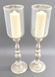 Elegante par de castiçais em metal prateado, acompanhado por cúpulas cilíndricas de vidro com friso metálico na borda; hastes de design clássico com refinado trabalho torneado, enriquecido por detalhe circular em resina translúcida. Acompanham duas velas brancas de espessura larga. Medidas: 57 x 15 cm.