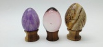 03 diferentes ovos decorativos, sendo dois em rochas lapidadas e polidas, e um em vidro; apoiados em bases individuais de madeira torneada. Medidas: 7 x 5 cm; 6 x 4 cm; 6 x 5 cm.