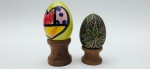 SILVANA PUJOL - 02 diferentes ovos decorativos, sendo um da artista brasileira Silvana Pujol (Pomerode, SC), em madeira pintada com motivos vegetalistas; e um em cerâmica esmaltada representando um coração, com cores fortes e vibrantes, assinado por artista não identificado; apoiados em bases individuais de madeira torneada. Medidas: 7 x 5 cm; 6 x 4 cm.