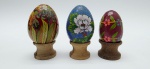 03 diferentes ovos decorativos, sendo dois em madeira pintados à mão com coloridos motivos florais e um em cerâmica azul, de temática oriental, com coloridos padrões florais e linhas em dourado; apoiados em bases individuais de madeira torneada. Medidas: 7 x 5 cm; 6 x 5 cm; 5 x 4 cm.