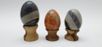 03 diferentes ovos decorativos em rochas lapidadas e polidas; apoiados em bases individuais de madeira torneada. Medidas: 6 x 5 cm; 5 x 4 cm. Uma das bases com ínfimo dano.