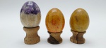 03 diferentes ovos decorativos em rochas lapidadas e polidas; apoiados em bases individuais de madeira torneada. Medidas: 6 x 4 cm; 5 x 4 cm.