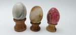 03 diferentes ovos decorativos em rochas lapidadas e polidas; apoiados em bases individuais de madeira torneada. Medidas: 6 x 5 cm; 5 x 4 cm. Ovo menor com rachaduras.
