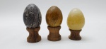 03 diferentes ovos decorativos em rochas lapidadas e polidas; apoiados em bases individuais de madeira torneada. Medidas: 6 x 5 cm; 5 x 4 cm.