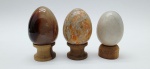 03 diferentes ovos decorativos em rochas lapidadas e polidas; apoiados em bases individuais de madeira torneada. Medidas: 6 x 5 cm; 5 x 4 cm.