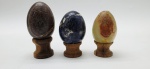 03 diferentes ovos decorativos em rochas lapidadas e polidas; apoiados em bases individuais de madeira torneada. Medidas: 6 x 5 cm; 5 x 4 cm. Um dos ovos com pontos trincados e ínfimo lascado.