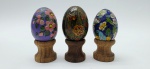 03 ovos decorativos com diferentes materiais e acabamentos, sendo um metal esmaltado com coloridos desenhos florais e borboletas em cloisonné; e um em cerâmica e um em madeira, com coloridos motivos florais e vegetalistas; apoiados em bases individuais de madeira torneada. Medidas: 6 x 5 cm; 5 x 4 cm.