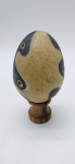 01 ovo decorativo artesanal em cerâmica, adornado com desenhos orgânicos nas cores bege e preta; com pequeno metal em seu interior agregando ao ovo a função de um suave instrumento de percussão; apoiado em base individual de madeira torneada. Medidas: 9 x 7 cm. Com ínfimos pontos de desgaste na pintura.