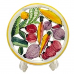 CERÂMICA LUIZ SALVADOR - Descanso de mesa adornado com coloridos legumes pintados à mão, filetes em amarelo e detalhes orgânicos vazados; em cerâmica esmaltada. Medidas: 25 x 3 cm.