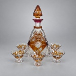 Conjunto para licor em cristal incolor lapidado, com reservas em tonalidade âmbar e decoração floral gravada, composto por uma licoreira com tampa e cinco taças. A licoreira apresenta corpo facetado, ornamentação com rosas e folhagens, gargalo com detalhe rosado e tampa facetada em cristal âmbar.A licoreira mede 27  11 cm, enquanto cada taça mede cerca de 5 cm de altura  7 cm de diâmetro. As taças repetem a decoração floral e os painéis âmbar da peça principal, formando conjunto harmonizado de seis peças. (lascado em um dos copos)