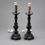 Par de antigos castiçais de altar executados em madeira entalhada e torneada, atualmente adaptados como bases de abajur. Medindo 75 x 25 cm cada. Apresentam bases trípodes recortadas, ornamentadas por volutas e elementos vegetalistas em relevo, fustes compostos por sucessivas reservas torneadas e entalhadas e pratos circulares superiores.