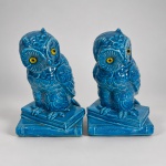 PAR DE ESCULTURAS DE CORUJAS  Executadas em cerâmica esmaltada medindo 18x12x11 cada em tom azul-turquesa, com olhos em amarelo e preto. As aves aparecem pousadas sobre bases moldadas em forma de livros empilhados, com plumagem trabalhada em relevo.