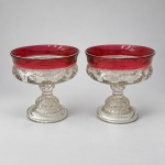 Par de elegantes taças em vidro moldado. Medindo: 13 x 13 cm cada. Apresentam copas largas e circulares, com bordas levemente abertas em tonalidade rubi, ornamentadas por relevos circulares e frisos serrilhados. As hastes moldadas repetem a decoração geométrica e repousam sobre bases circulares escalonadas.