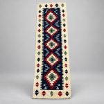 00969 Passadeira Kilim, confeccionada em lã sobre lã. Medindo: 2,72 x 0,78. campo central azul-marinho e moldura em tonalidade cru. Apresenta sequência de cinco medalhões escalonados em vermelho, verde e creme, intercalados por pequenos motivos estilizados. As bordas são decoradas com elementos geométricos repetidos nas mesmas tonalidades, formando composição simétrica.