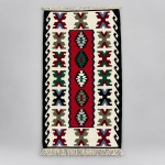 Tapete Kilim confeccionado em lã sobre lã com composição geométrica simétrica. Medindo: 1,26  0,67 m, apresenta campo central vermelho contendo cinco medalhões losangulares, ladeado por motivos escalonados sobre fundo creme e barras laterais pretas decoradas com elementos em forma de volutas.A composição emprega tonalidades de vermelho, verde, azul, preto, cinza e creme, com acabamento em franjas nas duas extremidades.