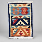 Tapete Kilim, confeccionado em lã sobre lã, peça com composição geométrica distribuída em três campos ornamentais. Medindo 54  83 cm, apresenta motivos escalonados e elementos abstratos em tonalidades de azul, terracota, vermelho, creme e amarelo.A composição é delimitada por moldura azul-marinho e faixa interna creme com pequenos motivos geométricos.