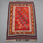 00421 TAPETE KILIM PERSA KASHIGAI  - manufatura, em lã sobre lã. Medidas: 2,82  1,63 m. Apresenta amplo campo vermelho ocupado por fileiras de medalhões geométricos, executados em marfim, azul-marinho, amarelo, laranja, verde, rosa e castanho. A composição é enquadrada por sucessivas bordaduras geométricas, destacando-se faixas em laranja e marfim, além de larga guarda externa formada por losangos escalonados em azul-marinho, branco, castanho e vermelho. Estado de conservação: apresenta desgastes de uso, áreas de abrasão, vincos, irregularidades e perdas nas franjas, além de reparos/restaurações visíveis junto às bordas e à união entre campo e moldura, conforme fotografias. ( Apresenta rugas nas bordas).