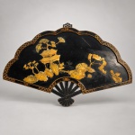 Painel decorativo de parede em formato de leque, confeccionado em madeira com acabamento laqueado preto e ornamentação dourada. Medindo 48 x 76 cm, apresenta composição central com paisagem oriental formada por pavilhões, árvores, rochedos e vegetação, executada com aplicações recortadas e gravadas em madrepérola de tonalidade dourada e iridescente. A moldura acompanha o contorno sinuoso do leque, com frisos dourados, arabescos e padrões geométricos repetidos. Na parte inferior, elemento vazado reproduz a armação de um leque aberto. Possui suporte metálico na parte superior para fixação na parede.