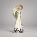 NADAL  Escultura decorativa espanhola modelo Sensaciones Mini Blanco (Feelings), pertencente à coleção Sirenes, executada em resina com acabamento marfim fosco e detalhes policromados. Medindo 17 x 7 x 6 cm, representa figura feminina de pé, trajando longo vestido de linhas fluidas e amplo chapéu, com formas alongadas e expressão serena. A barra e a lateral do vestido recebem decoração multicolorida em padrão de mosaico, característica recorrente da coleção e associada à linguagem ornamental inspirada nos mosaicos de Antoni Gaudí. Na base, apresenta inscrição original NADAL  No. 5235  Serie Numerada / Numbered Edition  Diseñado por / Designed by VALIENTE  Valencia - SPAIN