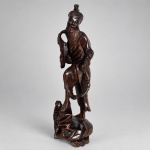 Escultura oriental em madeira escura entalhada e polida, representando personagem masculino barbado, trajando vestes longas e portando peixe e recipiente com alça. Medindo 30 x 9 cm, apresenta trabalho manual detalhado nas feições, barba, panejamento das vestes, escamas do peixe e elementos da base, que incorpora motivos vegetalistas e cesta.