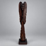 Escultura decorativa em madeira escura entalhada, representando figura angelical estilizada de corpo esguio, rosto geometrizado e asas verticais envolvendo a composição. Medindo 44 x 9 x 7 cm, apresenta acabamento polido, com entalhes marcando as penas das asas e o panejamento inferior, assentada sobre base quadrangular. Apresenta assinatura incisa na parte frontal da base.