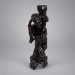 Escultura oriental em madeira escura entalhada e polida, representando personagem masculino de longas barbas, turbante e vestes amplas, disposto em composição vertical sobre base vazada em forma de rochedos. Medindo 31 x 12 x 10 cm, sustenta ao ombro grande recipiente de formato orgânico, elemento recorrente em representações chinesas de imortais portando cabaças e recipientes simbólicos. Apresenta rico trabalho de entalhe nas dobras das vestes, barba, cobertura da cabeça e base, com acabamento escuro lustroso. Na parte inferior conserva antiga etiqueta manuscrita.