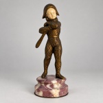 ARLEQUIM  Escultura decorativa de caráter Art Déco, executada em bronze patinado, representando personagem teatral trajando vestimenta com padrão losangular e chapéu bicorne. Medindo 19 x 7,5 cm, apresenta rosto esculpido em marfim, em contraste com o corpo em bronze de tonalidade dourada e escura. A figura segura um bastão junto ao corpo e repousa sobre base circular em mármore de coloração rosada e creme. O traje recebe decoração incisa em padrão geométrico, reforçando a identificação com a iconografia tradicional do Arlequim.