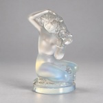 LALIQUE  Floreal, escultura francesa em cristal moldado, acetinado e opalescente, representando figura feminina nua ajoelhada, com um dos braços elevado sobre a cabeça e cabelos compostos por motivos florais. Medindo 8 x 5 cm, apresenta o contraste entre superfícies foscas e transparentes característico da manufatura Lalique. Criação de René Lalique, traz assinatura incisa Lalique France na base. O modelo Floreal foi concebido no século XX e posteriormente reeditado pela maison Lalique.