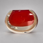 Peça de formato ovalado, executada em vidro artístico espesso incolor com núcleo interno vermelho, formando efeito sommerso. Medindo 13,5 x 18 cm, apresenta inclusões de bolhas alongadas distribuídas lateralmente, criando efeitos ópticos no interior da massa vítrea. A combinação entre o vermelho intenso, o vidro incolor e a construção maciça remete à produção decorativa italiana de meados do século XX.