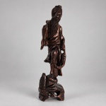 Escultura oriental em madeira maciça entalhada, representando pescador de longa barba, trajando vestes tradicionais e segurando peixe junto ao corpo. Medindo 31 x 9 x 9 cm, apresenta composição vertical sobre base trabalhada com elementos naturalistas, com detalhamento nas feições, barba, vestes e no peixe. A iconografia do pescador é recorrente na escultura decorativa chinesa, sendo frequentemente associada à abundância e prosperidade.