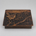 Caixa decorativa em madeira entalhada, de formato retangular, com tampa articulada ornamentada por cena em relevo representando ave pousada sobre galho, palmeiras e paisagem tropical. Medindo 5,5 x 20 x 14 cm, apresenta acabamento escuro com áreas ressaltadas em tonalidade castanha, frisos horizontais nas laterais e ferragens metálicas nas dobradiças. No interior da tampa, conserva inscrição entalhada S. SEBASTIÃO acompanhada de motivo geométrico.