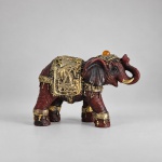 Escultura decorativa representando elefante asiático ricamente ornamentado, com corpo em tonalidade vermelho-escura e aplicações douradas. Medindo 6 x 8 x 3 cm, apresenta tromba erguida, presas claras, mantos e arreios trabalhados em relevo, além de pequeno elemento esférico alaranjado sobre a cabeça. A ornamentação metálica dourada recobre a manta, a cabeça e as patas, criando forte contraste com a coloração do corpo.