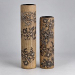 Vasos cilíndricos em cerâmica, com superfície externa texturizada em tonalidade bege e decoração floral em preto, formando ramos, flores e folhagens distribuídos verticalmente.O maior mede 40 x 10 cm e o menor 33 x 9 cm. Apresentam corpo alongado de linhas retas, textura fina que remete à trama de tecido e interior com acabamento preto brilhante, criando contraste com a superfície externa clara.