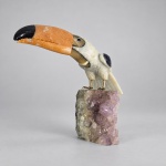 Escultura decorativa brasileira representando tucano, executada com diferentes pedras ornamentais entalhadas e polidas, montada sobre base natural de ametista. Medindo 25 x 30 x 16 cm, apresenta corpo em pedra clara, bico em tonalidade alaranjada com extremidade escura, detalhes das asas em pedra azul-violeta e olhos aplicados, com pés metálicos fixados diretamente sobre a drusa de ametista.