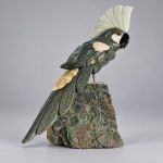Escultura decorativa representando cacatua, executada com diferentes pedras ornamentais entalhadas e polidas, montada sobre base natural de ametista. Medindo 29 x 21 x 15 cm, apresenta corpo em pedra verde escura, grande crista clara, bico em pedra preta, detalhes das asas em tonalidades rosadas e cauda alongada. A ave repousa sobre pés metálicos fixados diretamente sobre a drusa de ametista, cuja coloração violeta contrasta com as tonalidades da escultura.